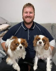 A smiling bearded man seated on a lounch with a Cavalier King Charles Spaniel on each knee.