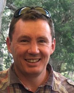 A smiling man with sunglasses perched on his head, wearing a brown and white checked shirt.