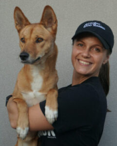 A smiling woman wearing a black cap and t-shirt is holding a tan and white dog in her arms.