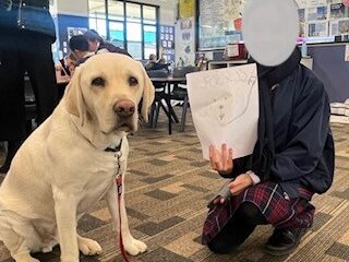 A yellow Labrador sitting on the floor beside a young girl who is holding up a picture she has drawn.