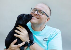 A smiling man wearing an aqua Guide Dogs t-shirt is holding a 6-week old black Labrador puppy which is licking his face.