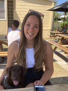 A smiling young woman is sitting at a picnic table with her arm around a chocolate Labrador puppy.