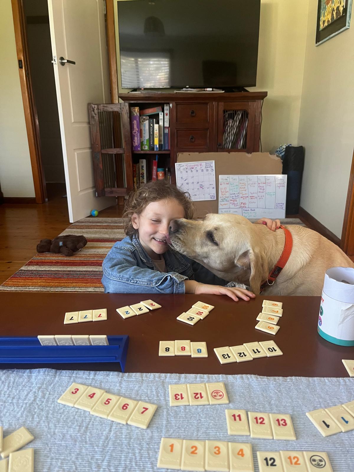 A yellow Labrador is licking the face of a young girl who is playing a tile game.